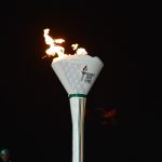 Prime plano da Tocha dos Jogos da Juventude da Região 5 (AUSC) acesa contra um fundo preto, exibindo o logótipo oficial e as cores das nações da África Austral.