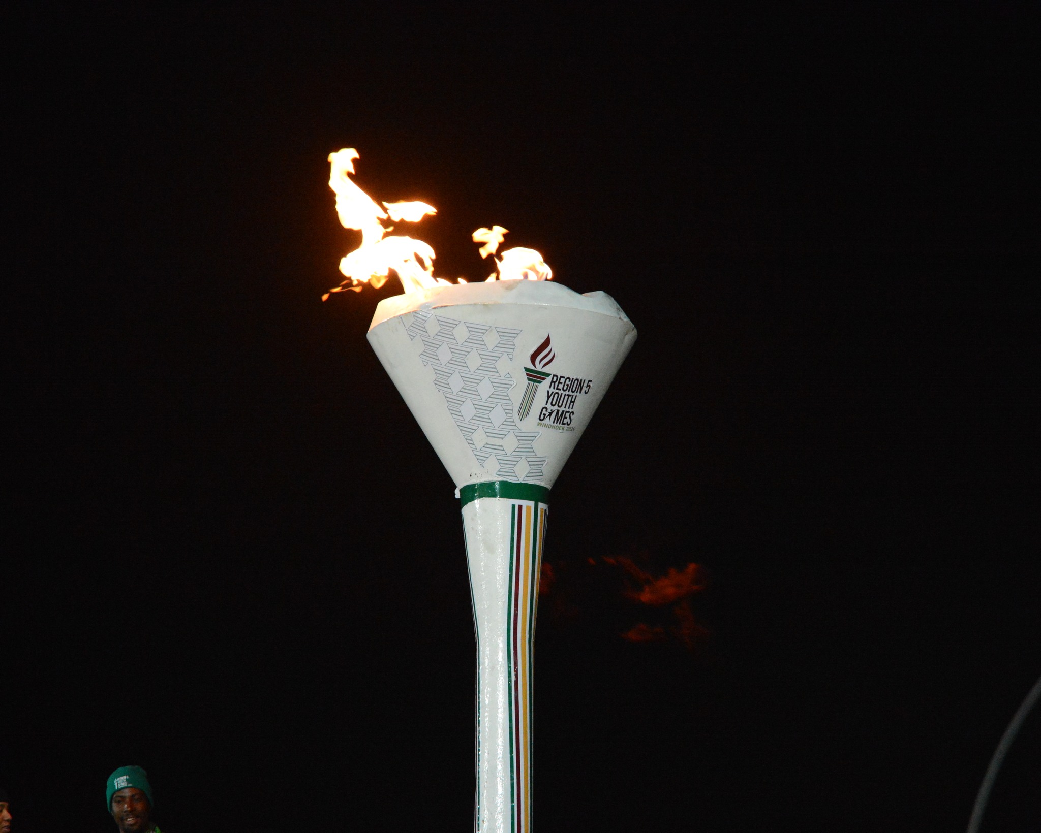 Prime plano da Tocha dos Jogos da Juventude da Região 5 (AUSC) acesa contra um fundo preto, exibindo o logótipo oficial e as cores das nações da África Austral.