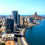 Vista aérea da orla de Dakar (Corniche) com edifícios modernos, o mar e o Monumento à Renascença Africana ao fundo sob céu limpo.