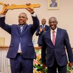 O Ministro Eugénio Laborinho segura uma grande chave simbólica de madeira acima da cabeça, ao lado de outra figura governamental em traje formal. Ambos sorriem durante uma cerimónia oficial num auditório decorado com flores.