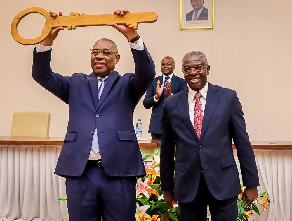 O Ministro Eugénio Laborinho segura uma grande chave simbólica de madeira acima da cabeça, ao lado de outra figura governamental em traje formal. Ambos sorriem durante uma cerimónia oficial num auditório decorado com flores.
