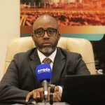 Retrato em grande plano do Secretário de Estado para as Finanças e Tesouro de Angola, Ottoniel dos Santos. Ele veste um fato cinzento escuro, camisa branca e gravata, usa óculos de armação preta e está sentado atrás de um microfone azul da agência ANGOP. À sua frente encontra-se um computador portátil.