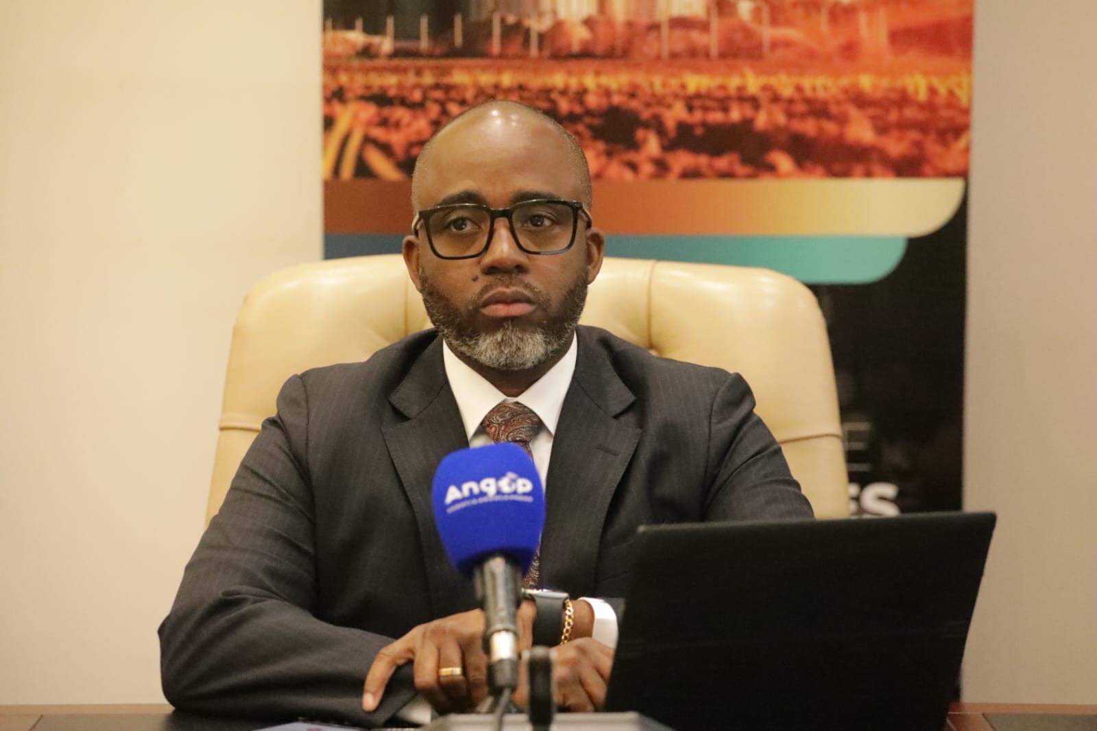 Retrato em grande plano do Secretário de Estado para as Finanças e Tesouro de Angola, Ottoniel dos Santos. Ele veste um fato cinzento escuro, camisa branca e gravata, usa óculos de armação preta e está sentado atrás de um microfone azul da agência ANGOP. À sua frente encontra-se um computador portátil.